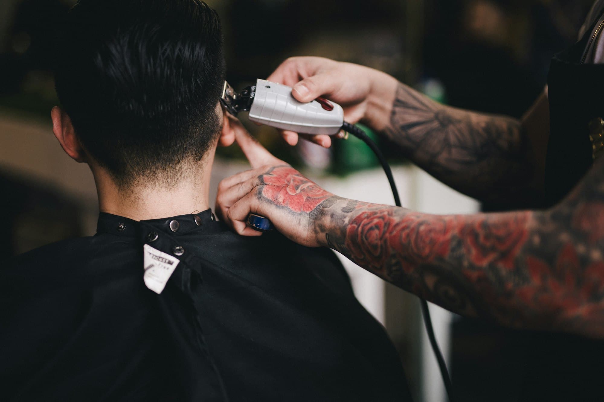 A barber with tattooed forearms running clippers along the neckline of a man seen from behind, wrapped in a black cape
