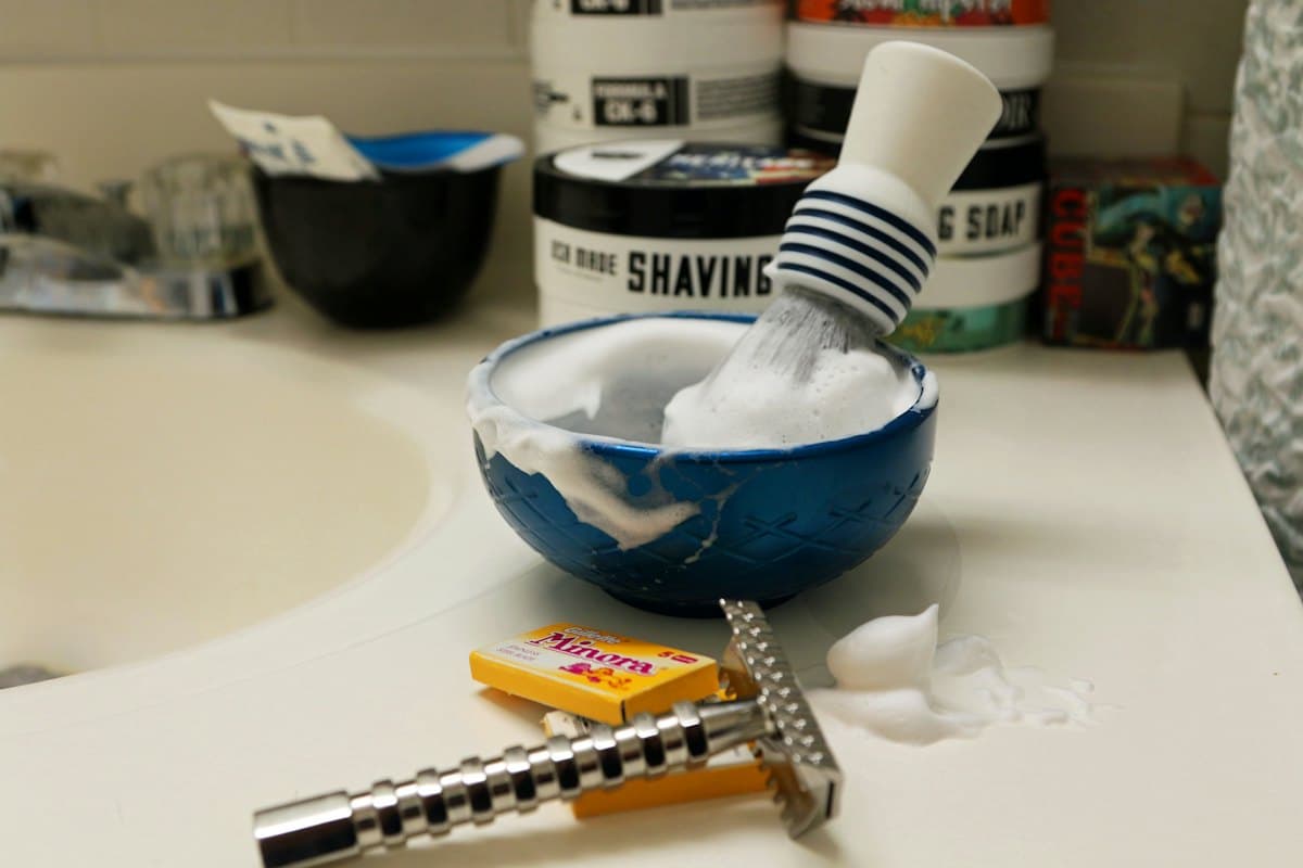 A shaving brush standing in a bowl of lather beside a safety razor