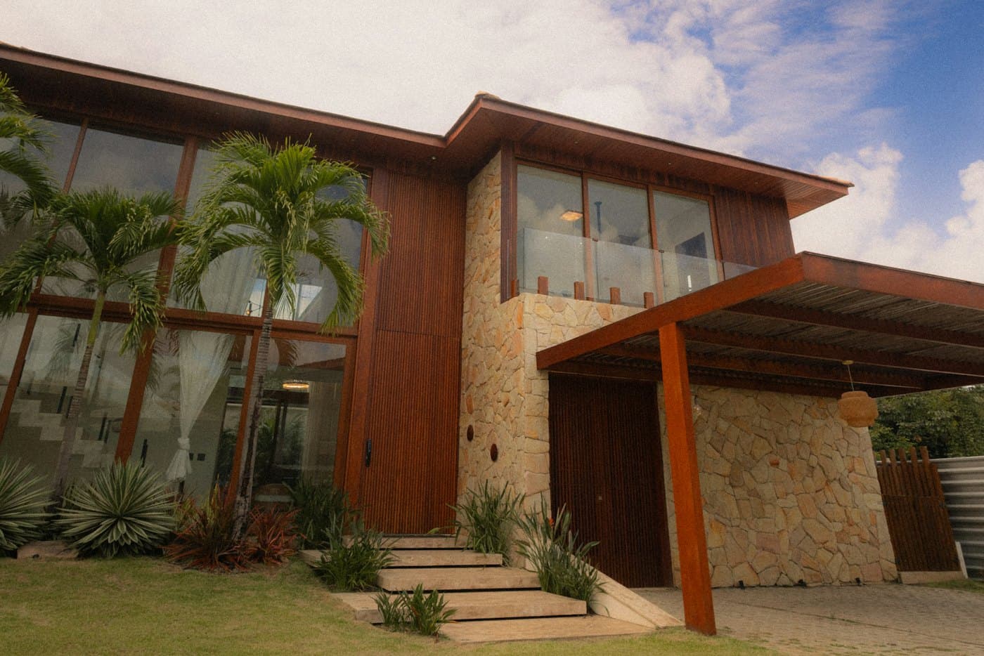 Corner of a timber-clad two-storey house with palms and a stone wall
