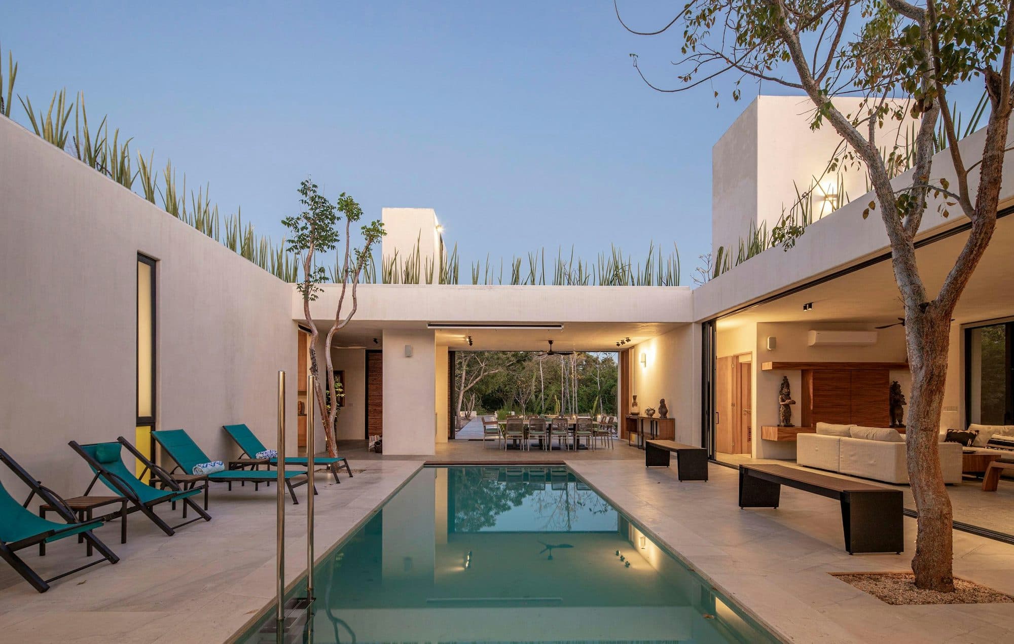 A white courtyard house at dusk: a long pool runs between a covered dining terrace and an open, lit living room