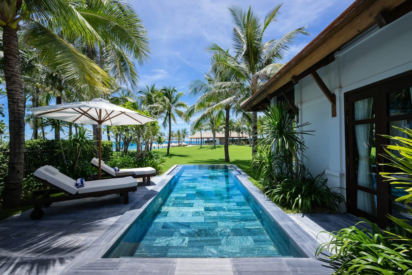 Long green stone lap pool beside a white house and palms