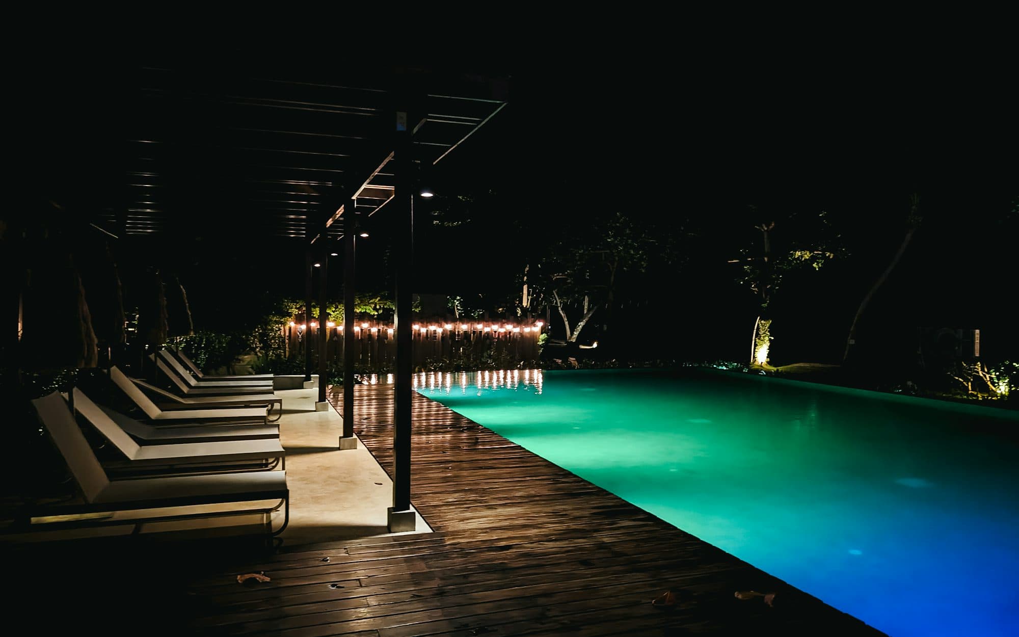 A long swimming pool glowing green in the dark, beside a wooden deck, loungers and a string of small lights