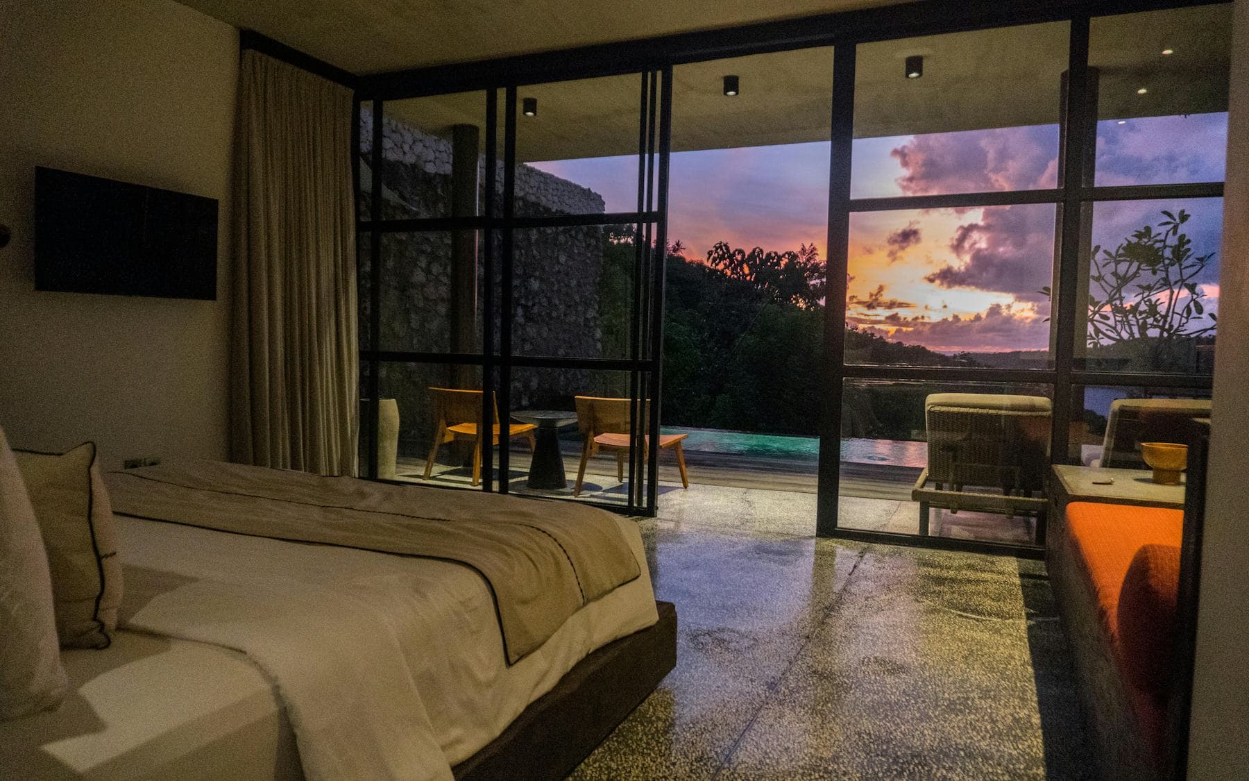 A bedroom at dusk with a glass wall open to a private pool terrace and a violet and orange sky
