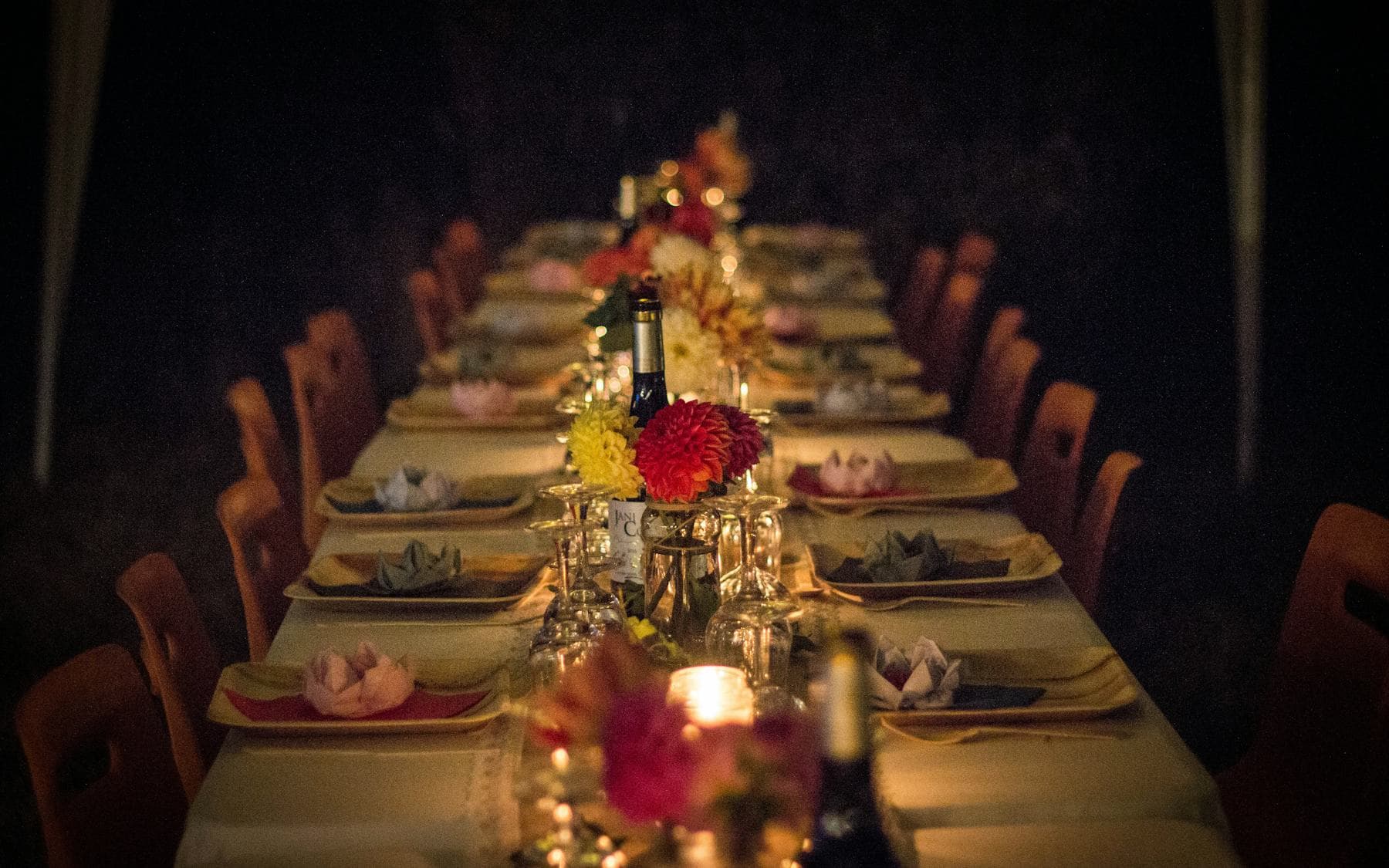 A long dining table in the dark, set for many guests and lit only by candles and flowers down its centre