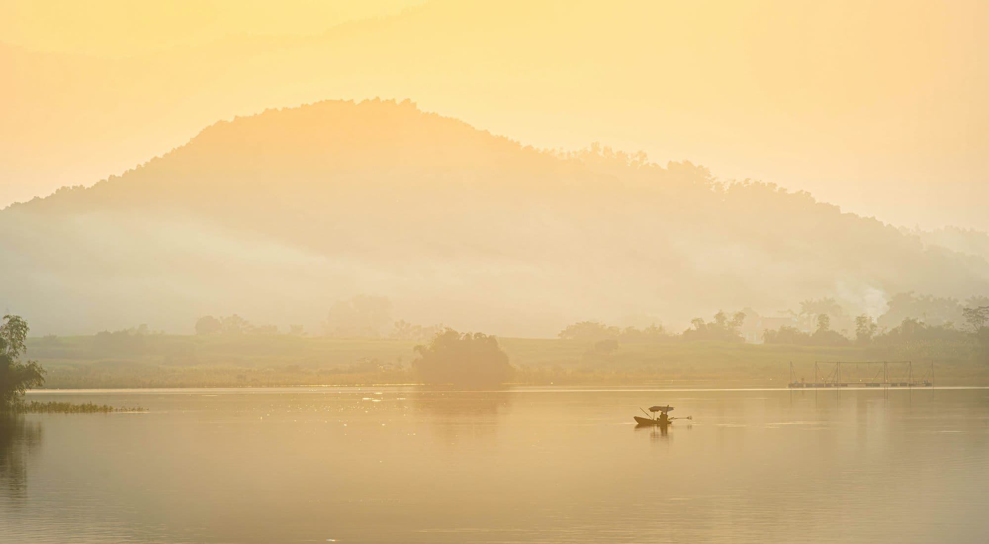 A lake in golden haze at sunrise, a lone boat on the water and a mountain fading into mist behind