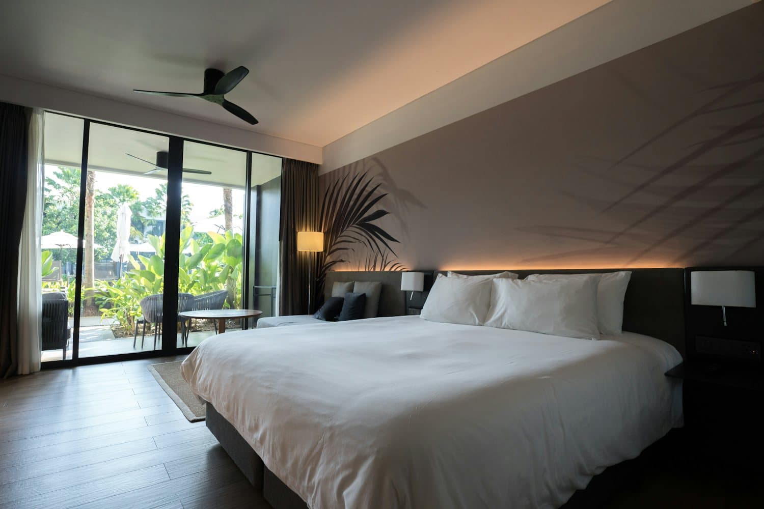 A calm, dim bedroom with a wide white bed and floor-to-ceiling glass doors opening onto a terrace and trees