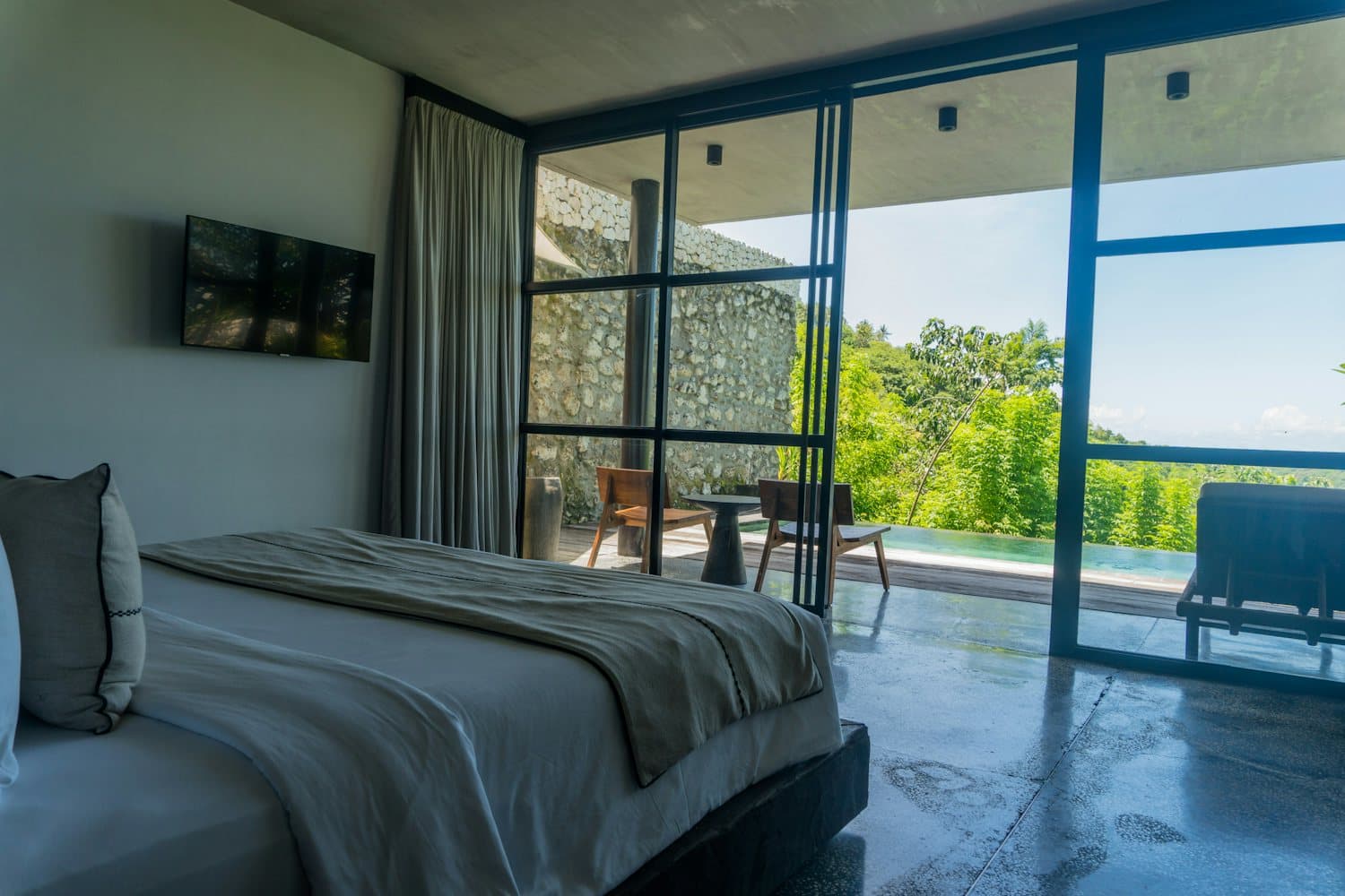 A bedroom with a polished stone floor and a steel-framed glass wall opening to a private pool and green hills