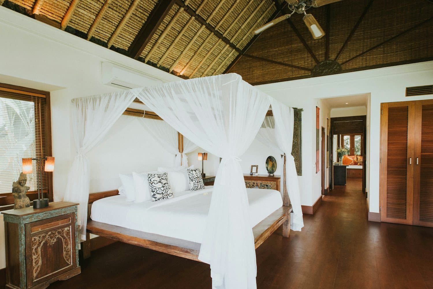 A white canopy bed draped in muslin beneath a high thatched roof, with carved wooden furniture and a long hall beyond
