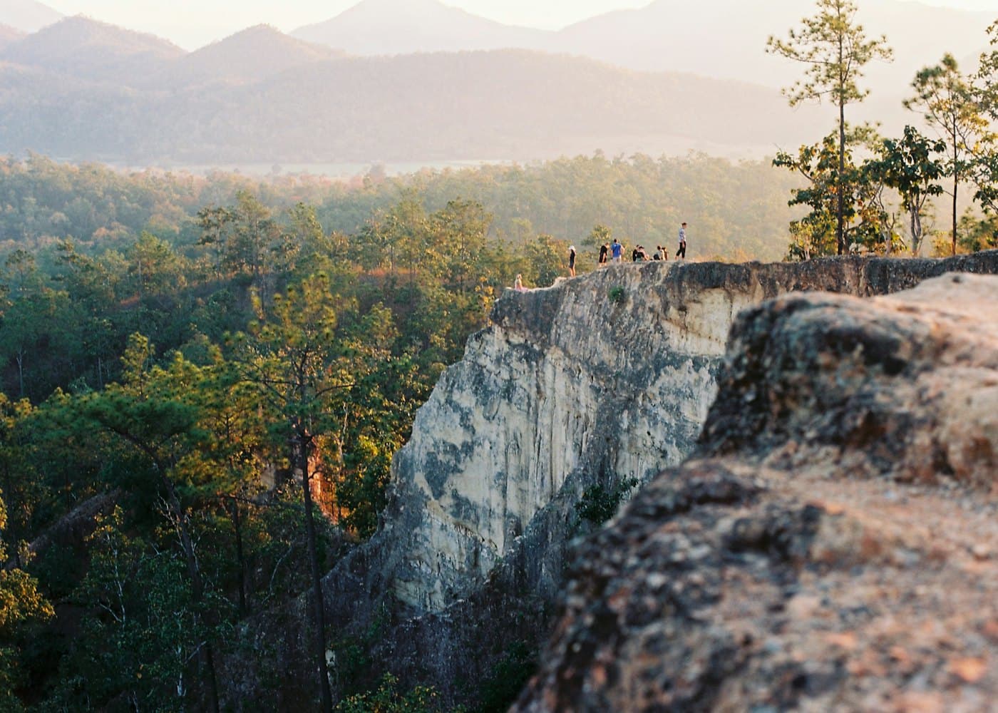 A pale sandstone ridge with a few people standing on its edge above pine forest, hills fading behind in evening light