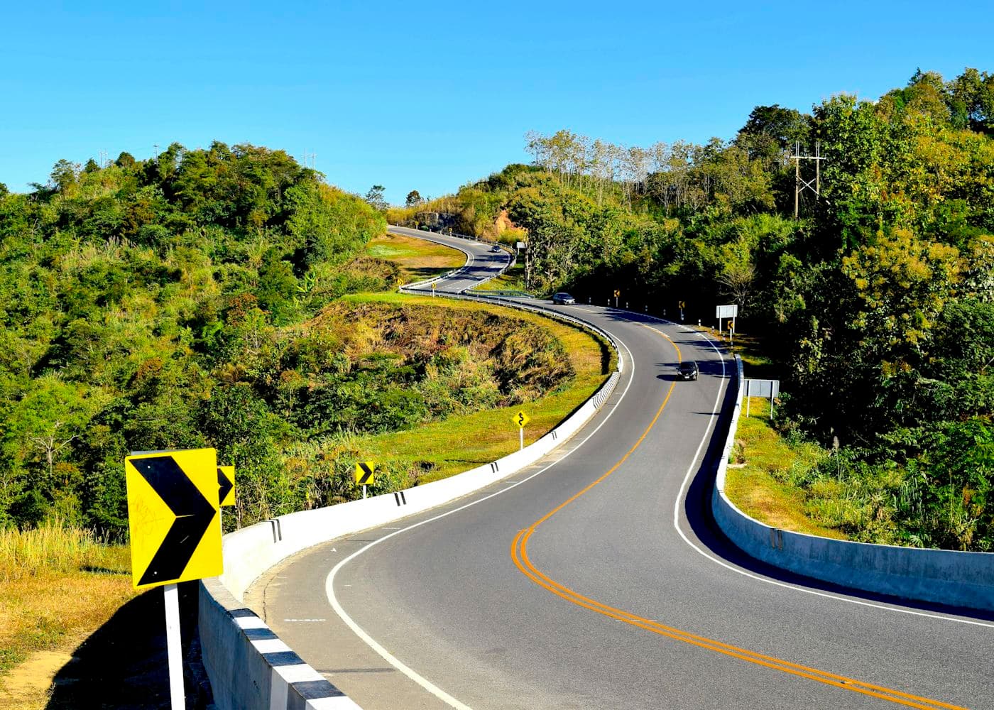 A freshly surfaced mountain road sweeping through an S-bend past yellow and black chevron signs under a blue sky