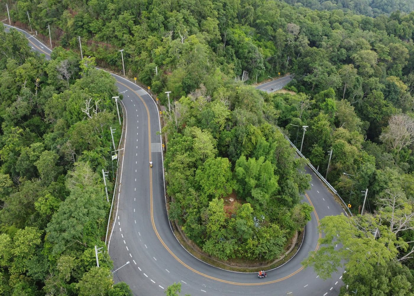 A hairpin bend in a two-lane mountain road seen from above, wrapped in dense green forest