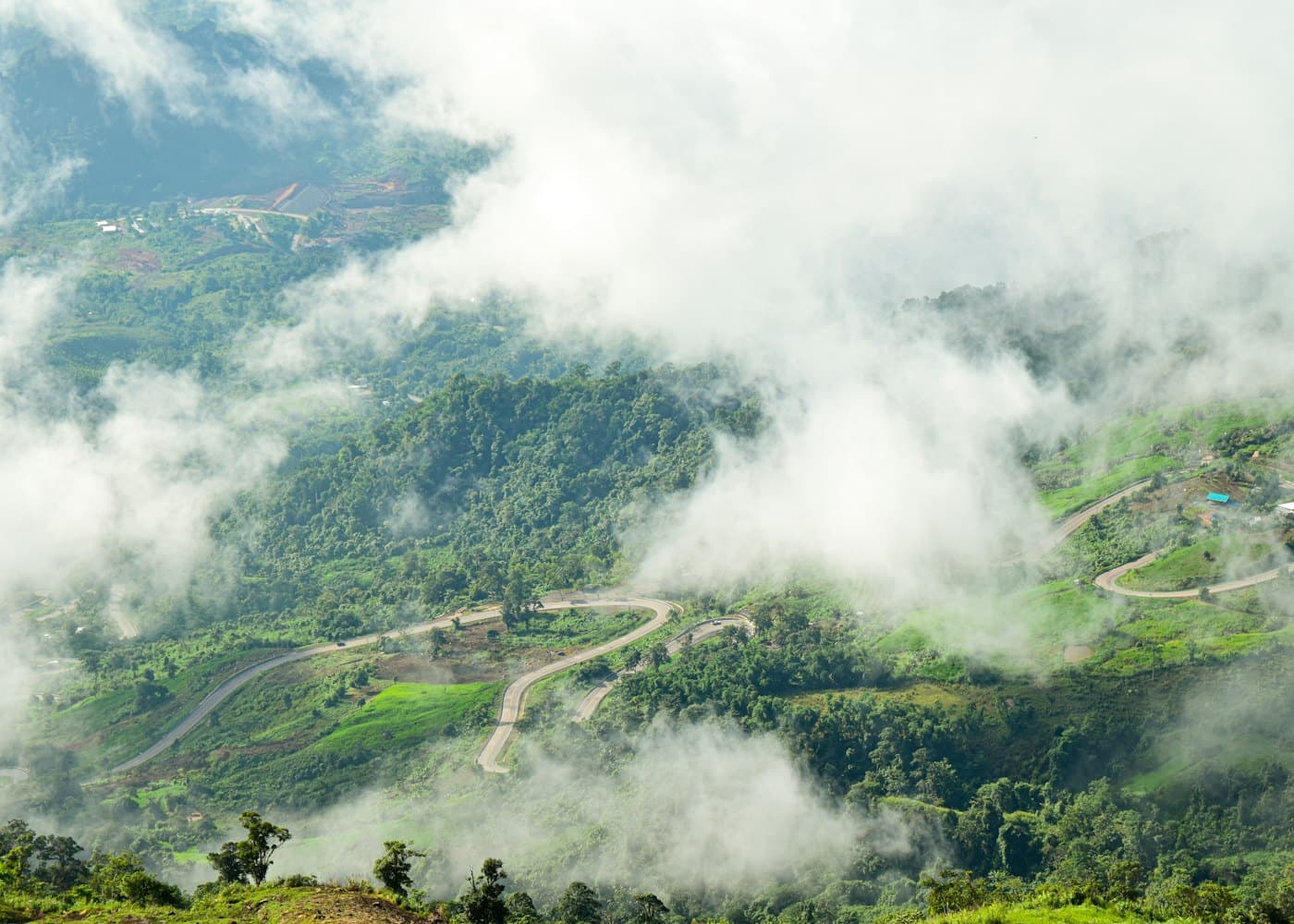 A narrow road winding along green terraced hillsides far below, half hidden under low white cloud