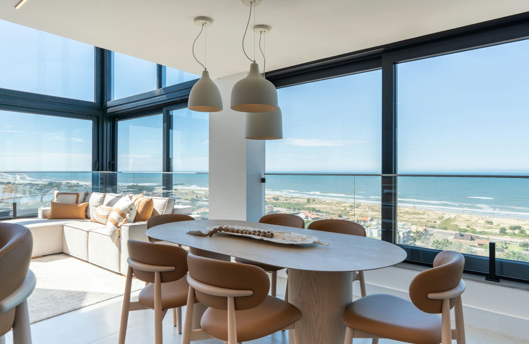 A dining table and tan chairs in a glass-walled corner room high above a long sandy beach