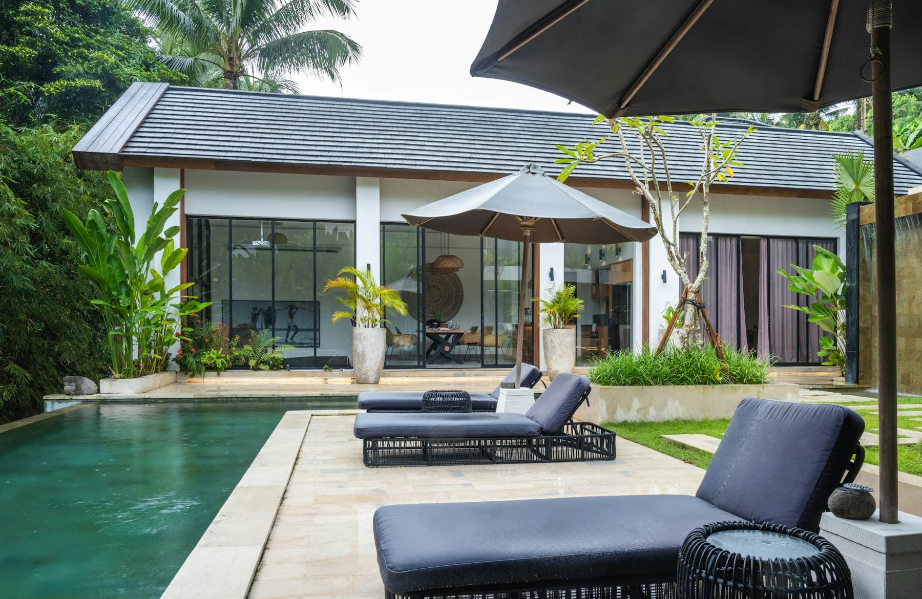 A single-storey villa with dark-framed glass doors facing a green pool, sun loungers and a parasol