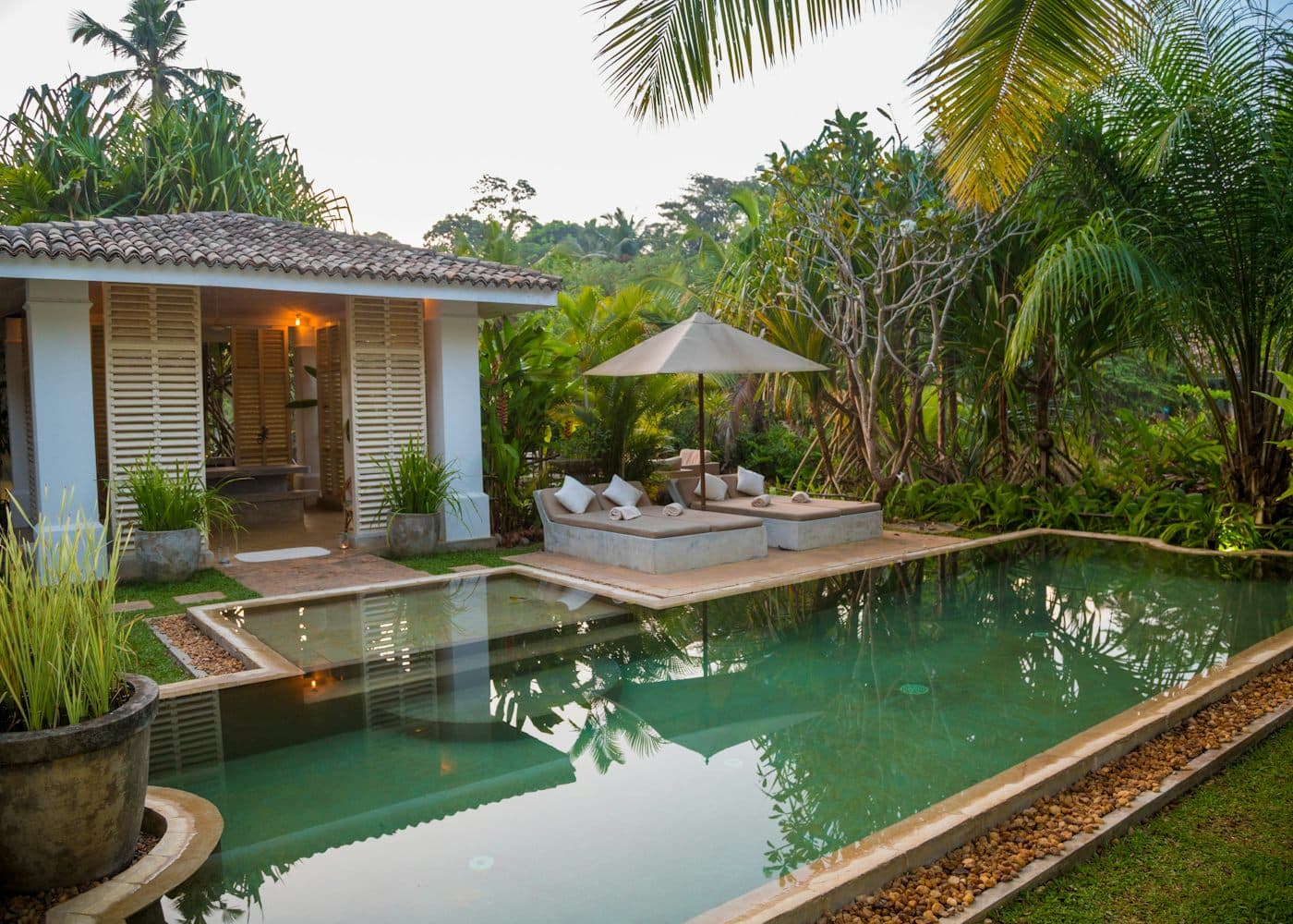 A white house with louvred shutters beside a long green pool, surrounded by palms