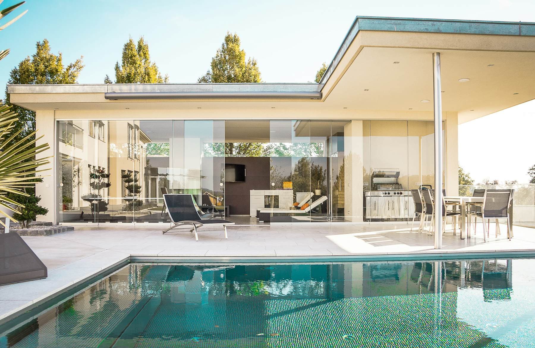A low modern house with floor-to-ceiling glass walls beside a swimming pool in bright sun