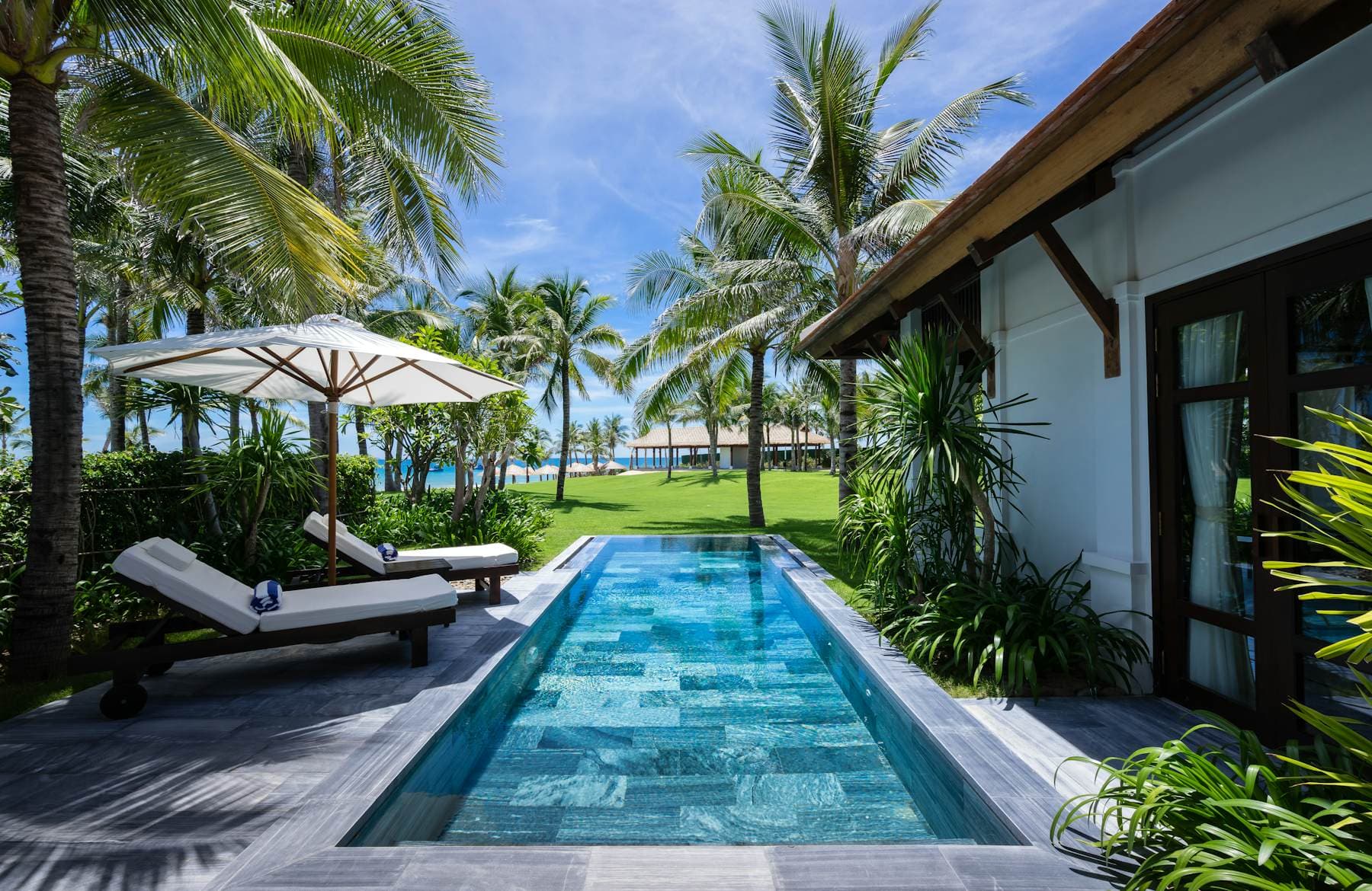 A long slate lap pool beside a white villa, with sun loungers, parasols and coconut palms on a lawn