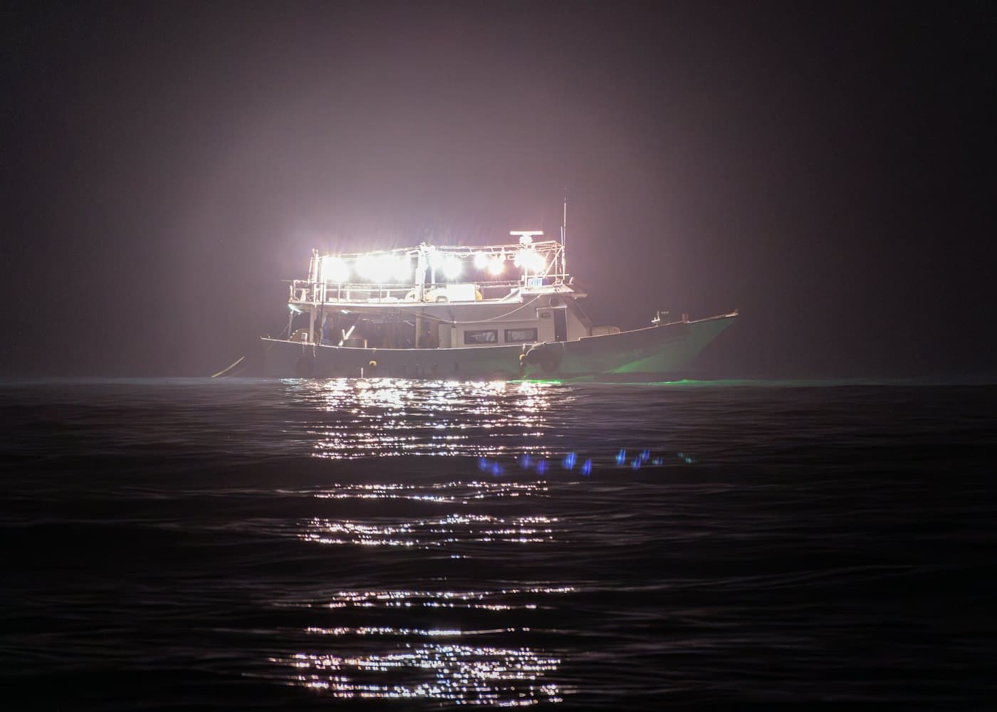 A squid boat at night, lamps blazing, a green glow on the water beside its hull