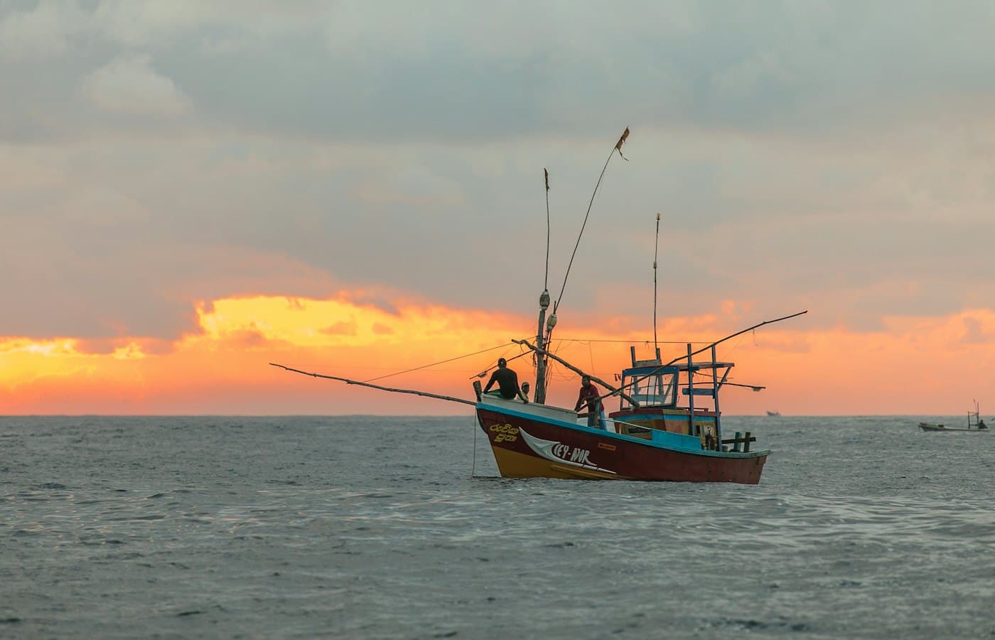 A small fishing boat with two men aboard crossing grey water under an orange sunset