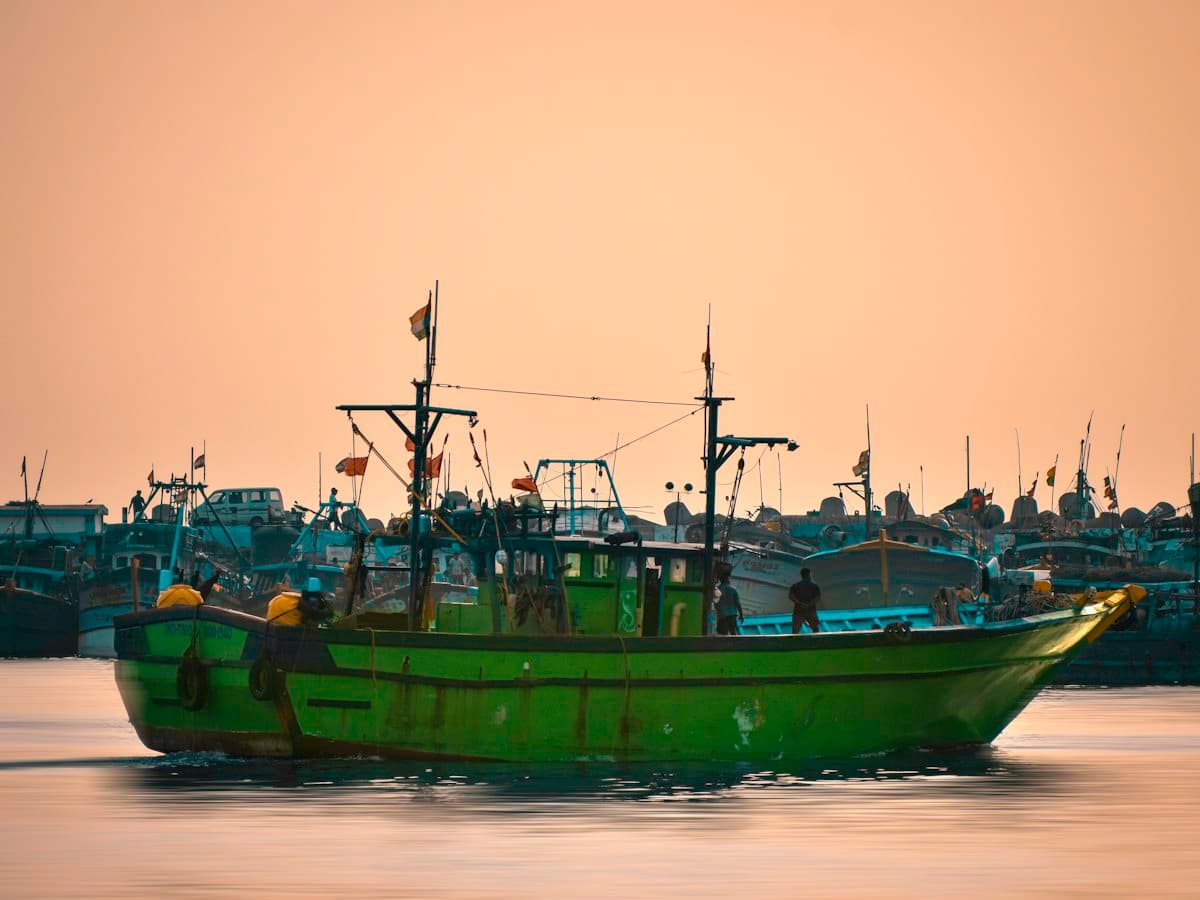 A green wooden fishing boat crossing a harbour under a peach evening sky