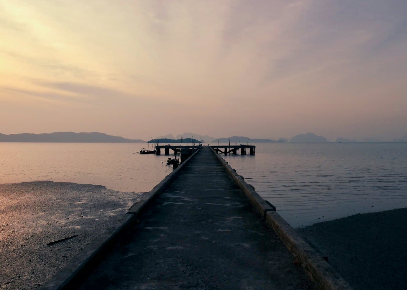 A long concrete pier running out into a calm sea under a pink and gold sky, islands on the horizon