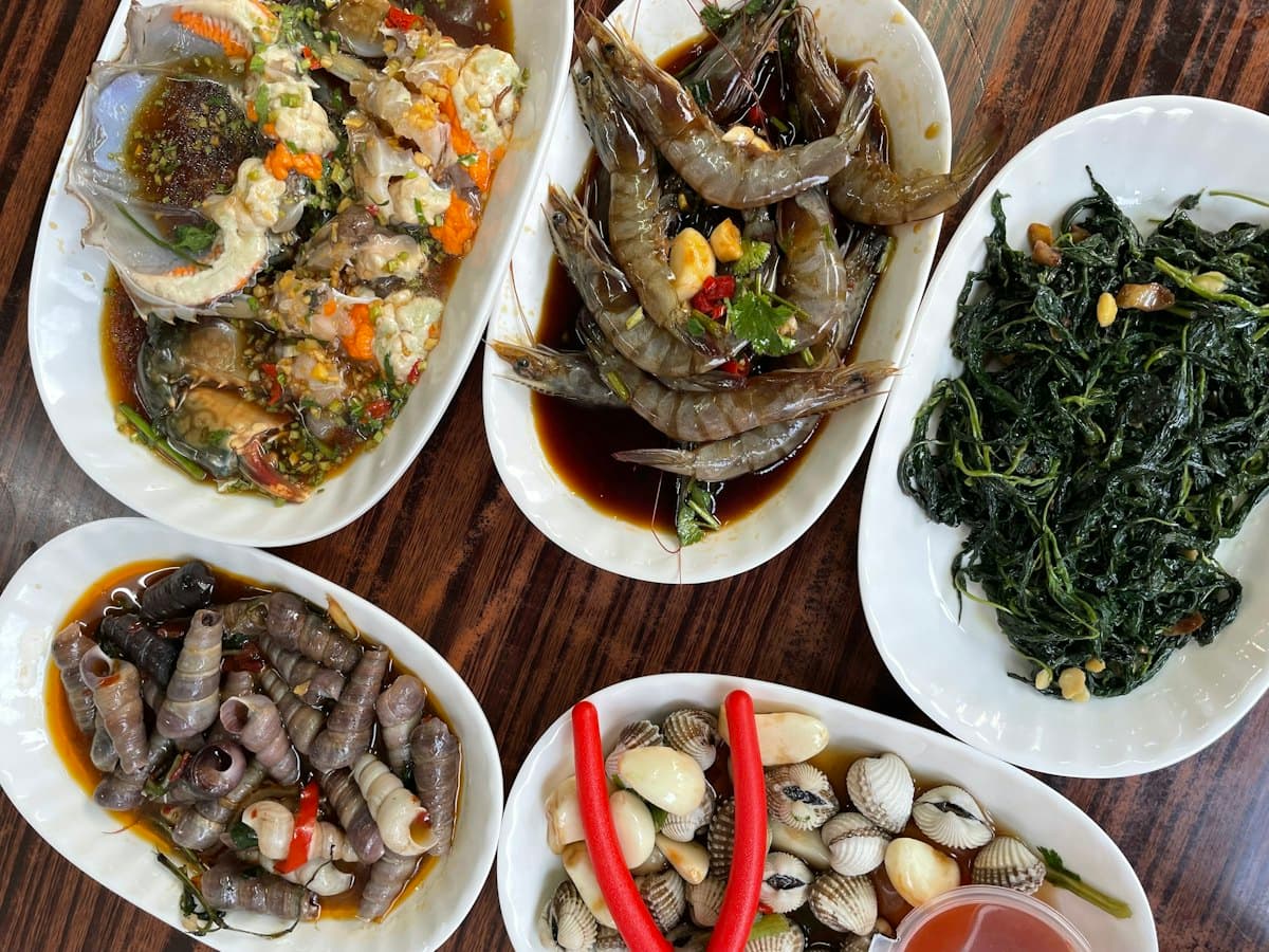 Five white plates on a dark wooden table: marinated crab, prawns, sea snails, clams and stir-fried greens