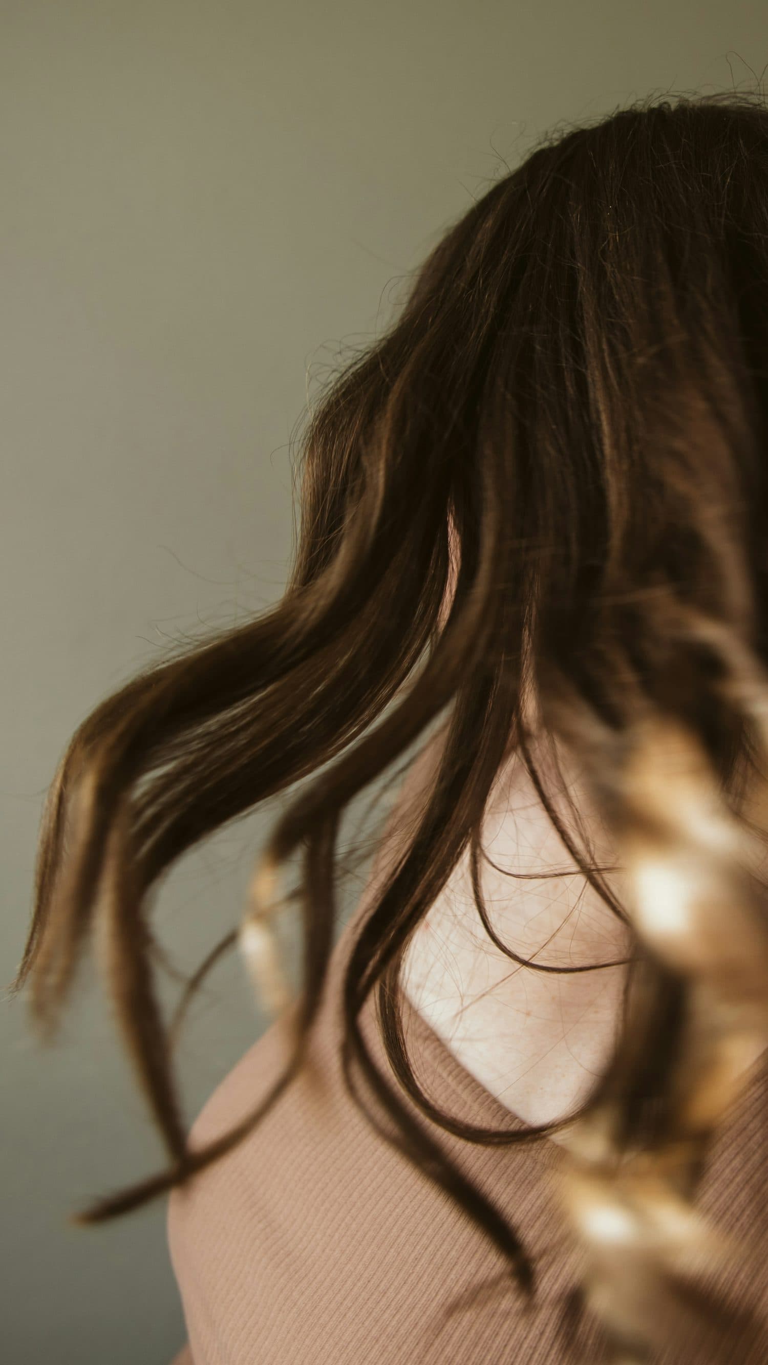 Long dark-brown hair with soft golden ends, caught mid-swing over a shoulder against a calm grey-green wall