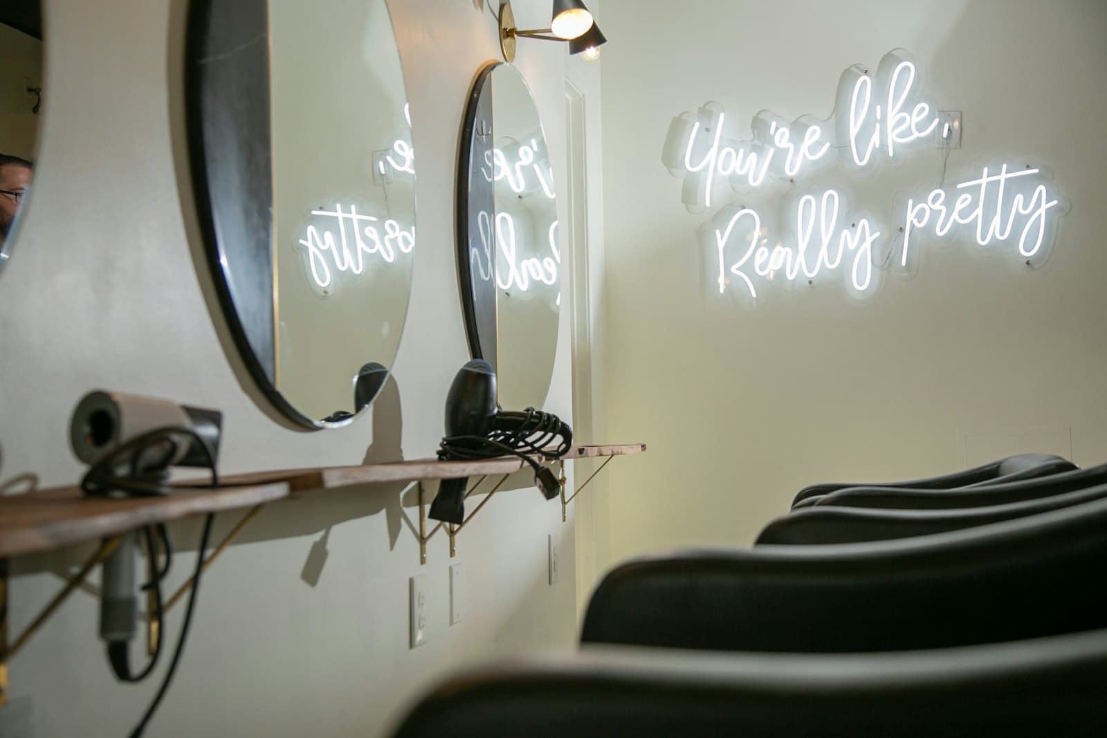 Round mirrors on a pale wall above a wooden shelf and dark salon chairs, with a soft white neon sign glowing beside them