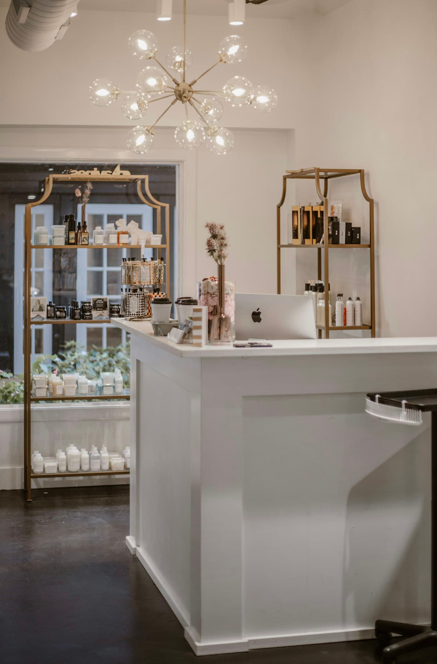 A white reception counter with gold shelving behind it, stocked with bottles and a small plant