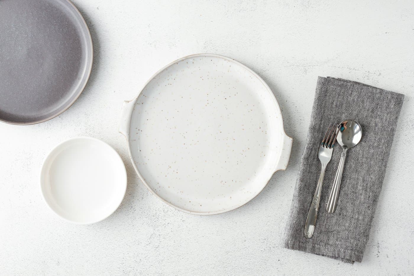 A speckled white handled plate, a small white dish and a grey plate laid on a table with a linen napkin, fork and spoon