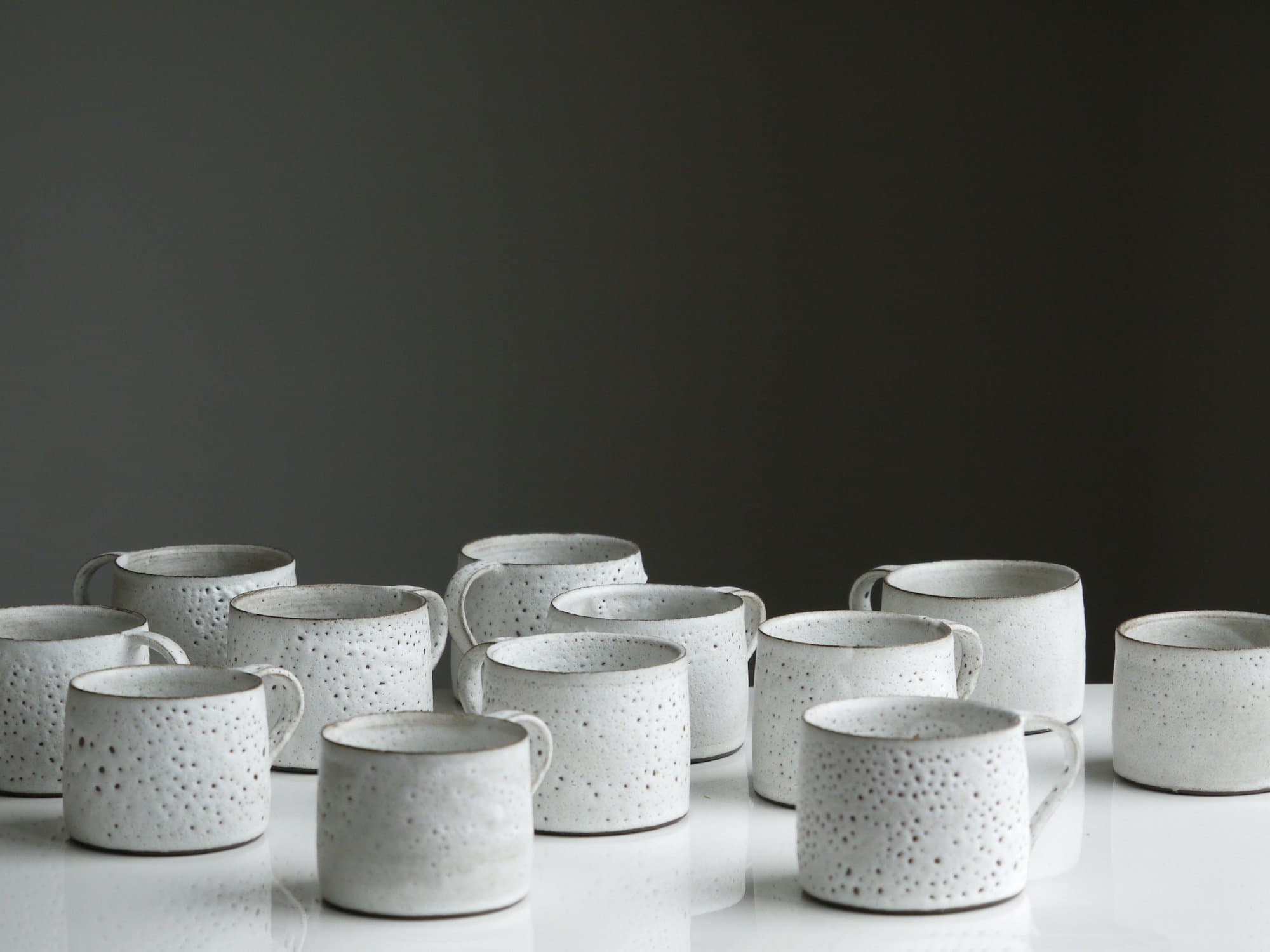 A dozen hand-thrown white mugs with a pitted crater glaze, spread across a white table in front of a dark wall