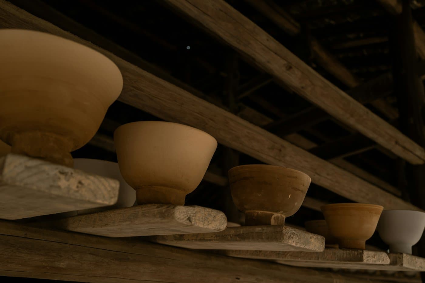 Unfired clay bowls drying on rough wooden planks under the dark beams of a barn roof