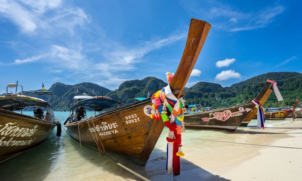 Longtail boats on a white beach, their tall prows wrapped in coloured ribbons and flower garlands