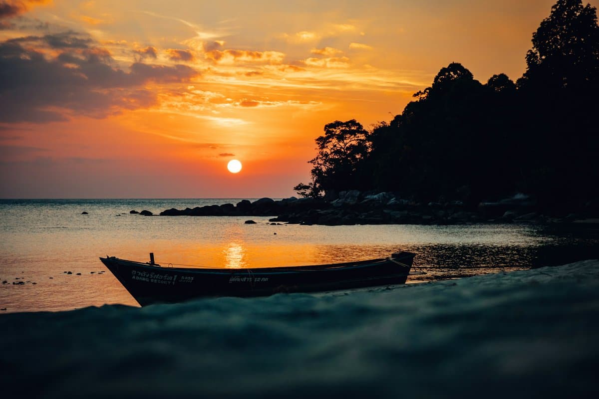 A small boat anchored off a dark shore under an orange sunset, with islands on the horizon