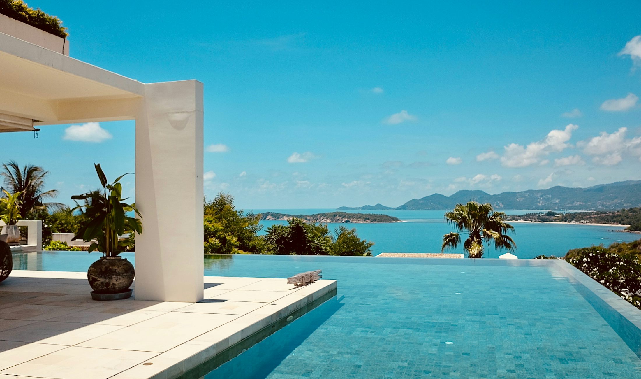 An infinity pool on a white villa terrace, looking over palms to a blue bay and the hills of Koh Samui